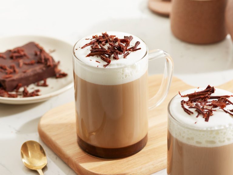 Một cốc thủy tinh chứa cà phê mocha với lớp kem sữa và vụn sô cô la đen, đi kèm với một đĩa bánh tráng miệng.
