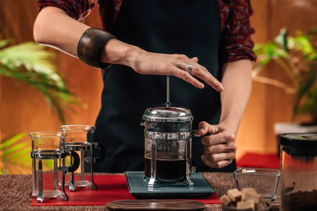 Barista pressing down the plunger on a French Press with two glass mugs beside them.