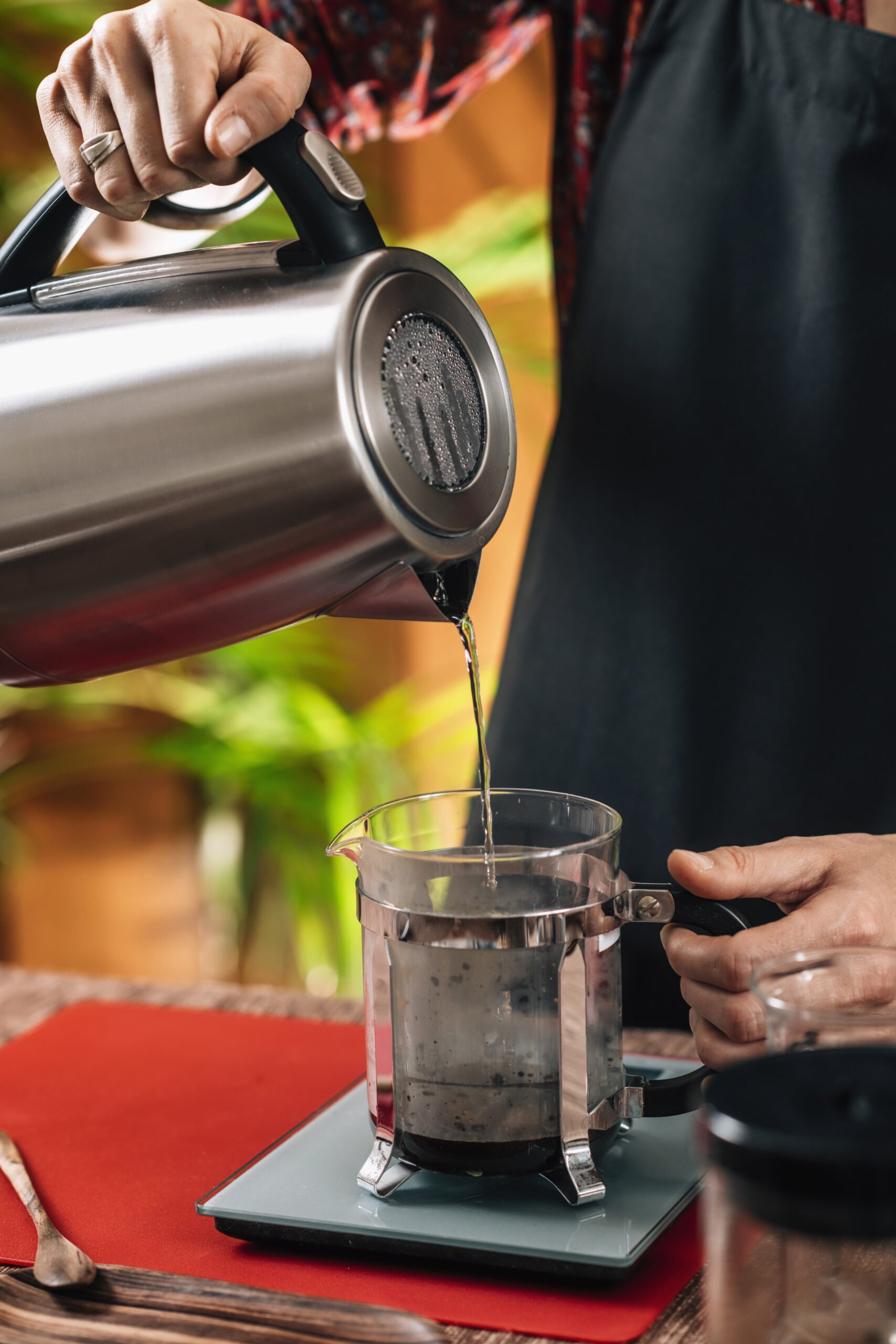A person pours hot water from a kettle into a French press filled with coffee grounds.