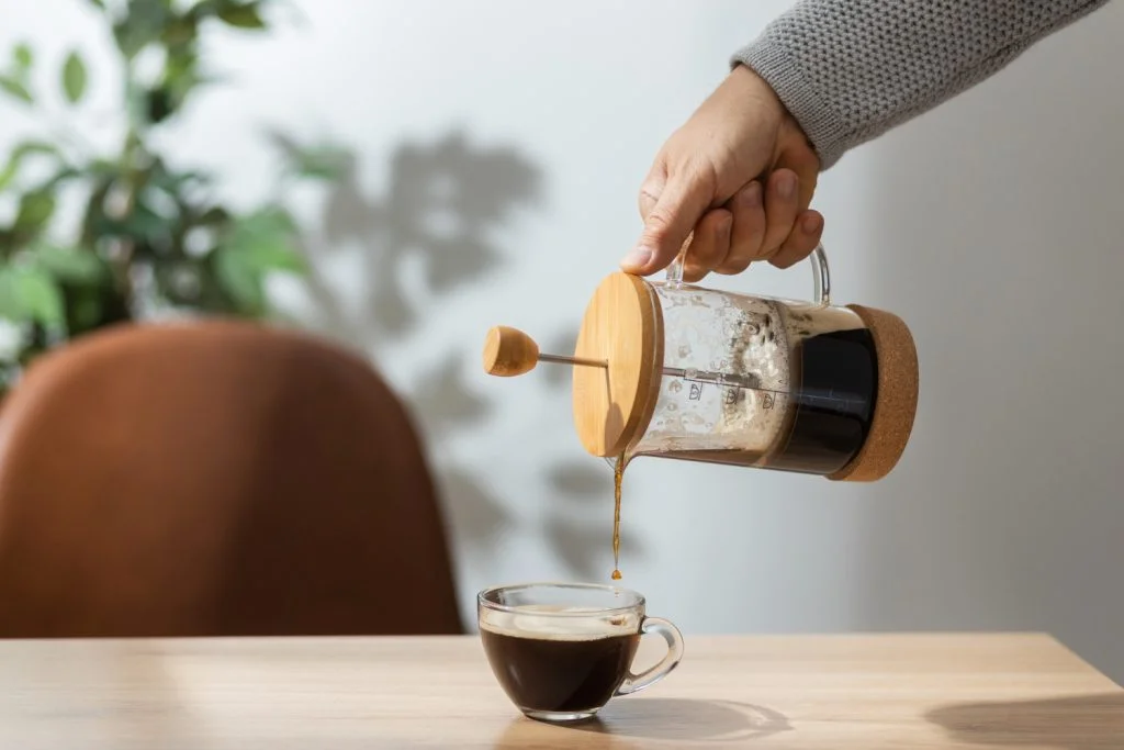 A hand pouring coffee from a French press into a glass cup on a wooden table.