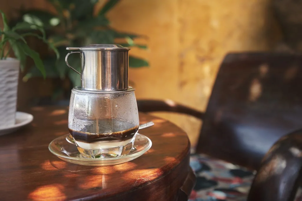Vietnamese phin drip coffee filter brewing atop a glass, with a cozy café ambiance.