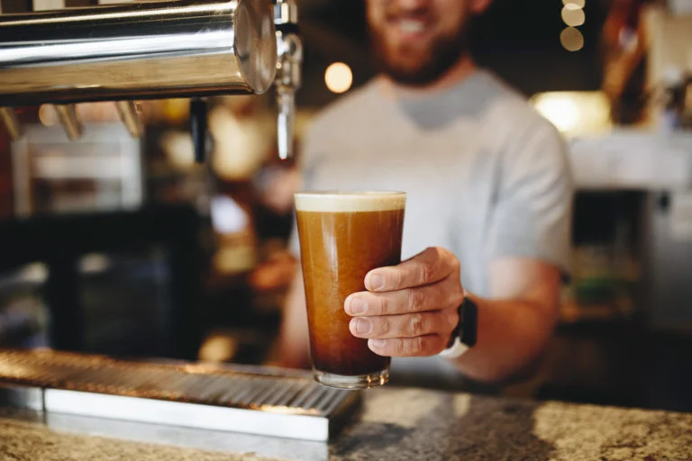 A barista smiles as he holds a freshly poured glass of nitro cold brew coffee.