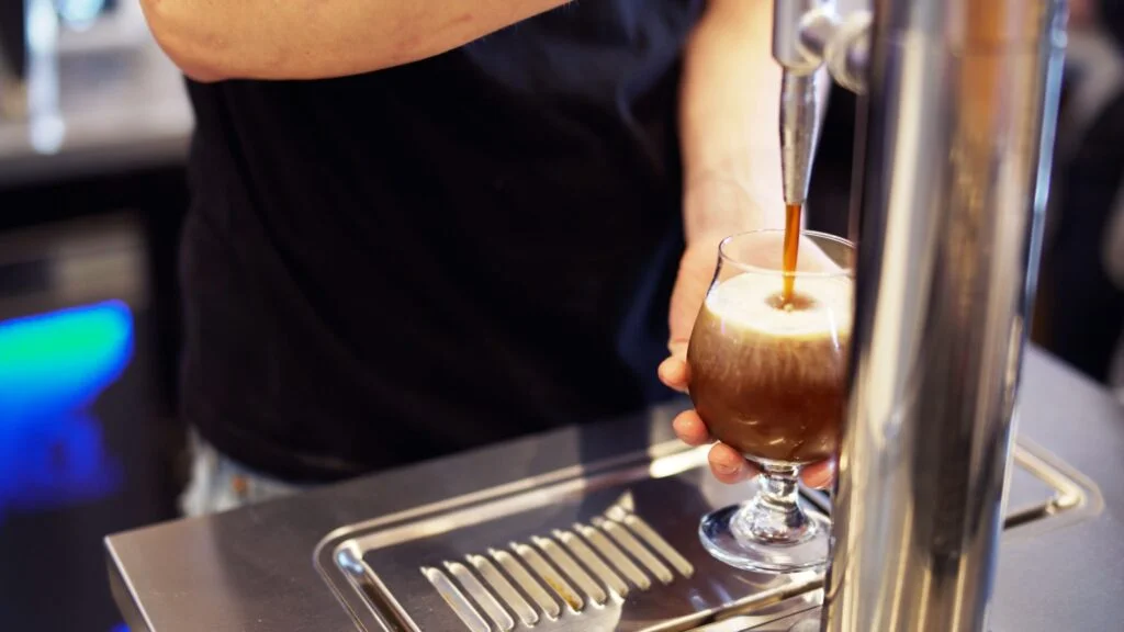 A barista pours nitro cold brew coffee from a tap, creating a cascading effect in the glass.
