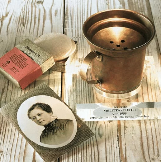 The first Melitta coffee filter from 1908, invented by Melitta Bentz, displayed with a portrait of Melitta Bentz and original packaging.
