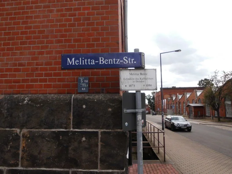 Street sign for Melitta-Bentz-Straße on a brick building corner, with a commemorative plaque for Melitta Bentz.