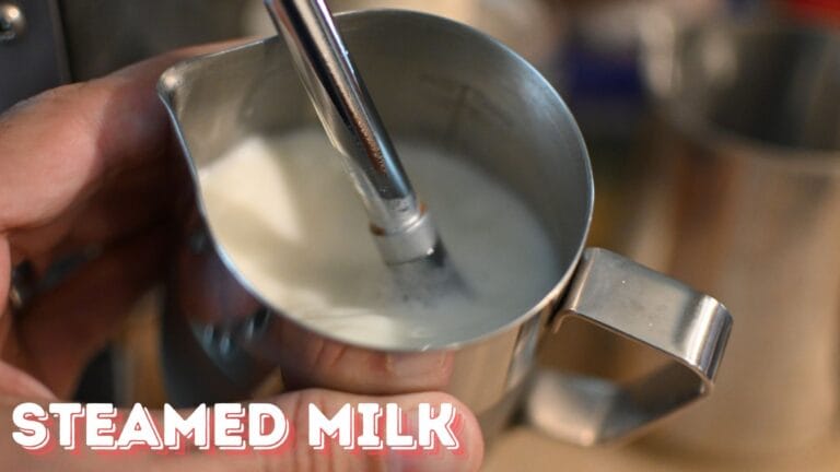 Close-up shot of a professional steam wand aerating milk in a stainless steel pitcher, creating microfoam for specialty coffee drinks. Steam rises from the surface as the milk is being textured.