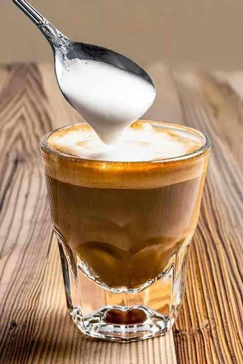 A silver spoon pouring steamed milk foam over a layered cortado coffee in a clear glass on a wooden table