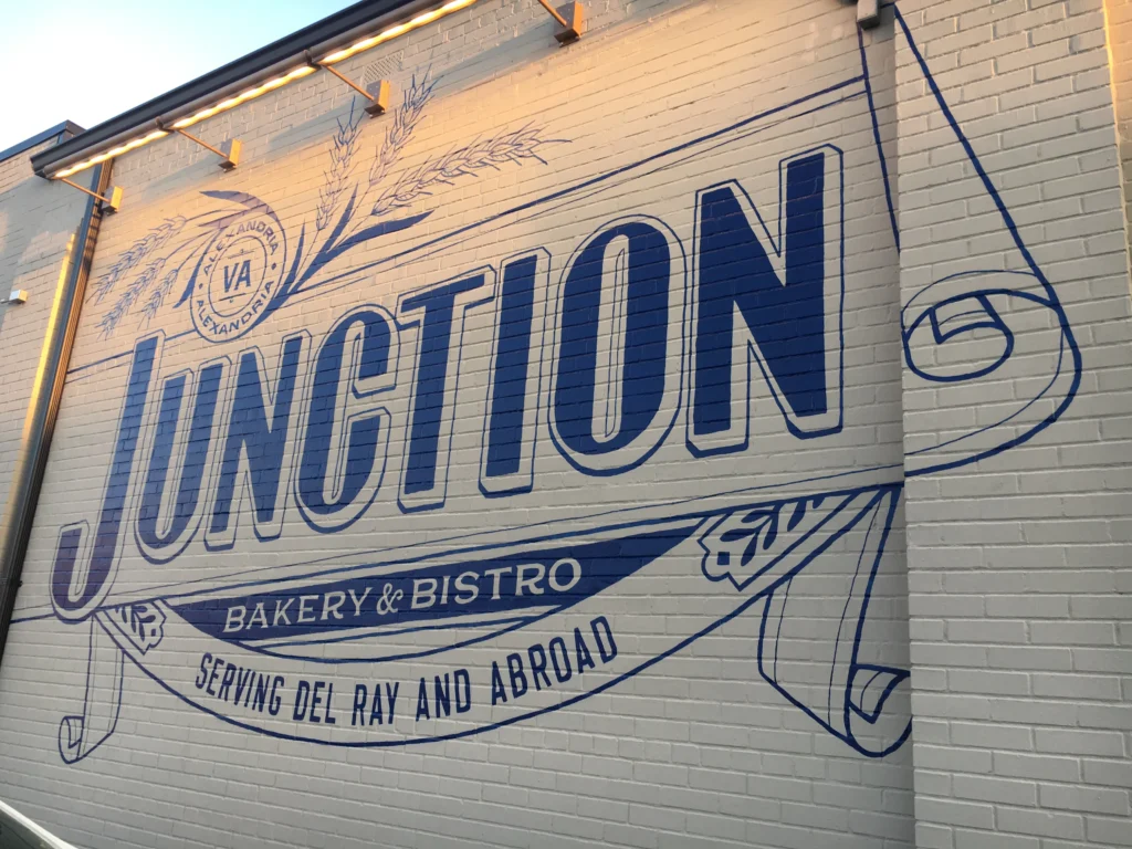 Alexandria's Ultimate Cortado Cafe & Coffee Shop Guide 2025 11 Large navy blue mural on white brick wall featuring Junction Bakery & Bistro's logo, decorative wheat stalks, VA seal, and "Serving Del Ray and Abroad" tagline, illuminated by overhead lighting.