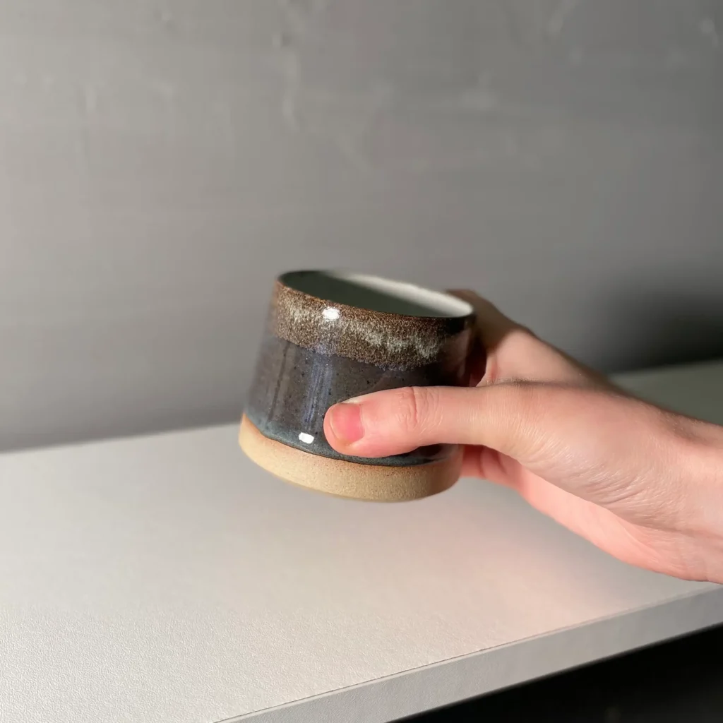 A hand holding a Fourth Ceramics Black Pearl handmade ceramic cup, showcasing its unique reactive glaze for flat white cups.