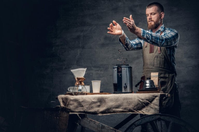 Bearded barista in apron carefully pouring coffee beans for 3rd wave coffee brewing with Chemex and specialty equipment on rustic table