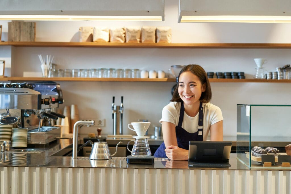 Smiling female 3rd wave barista behind modern coffee shop counter with pour over equipment, gooseneck kettle, and specialty coffee bags on minimalist wooden shelves
