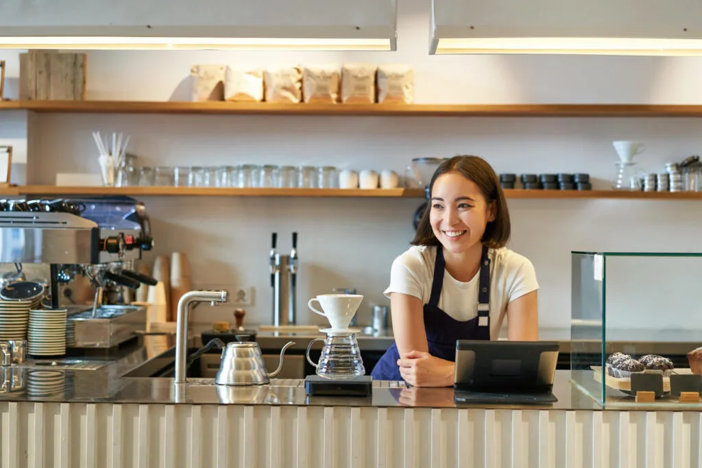 Smiling female 3rd wave barista behind modern coffee shop counter with pour over equipment, gooseneck kettle, and specialty coffee bags on minimalist wooden shelves