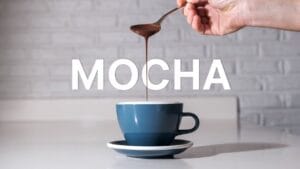 Hand drizzling chocolate syrup into blue coffee cup showing origins of the mocha drink preparation