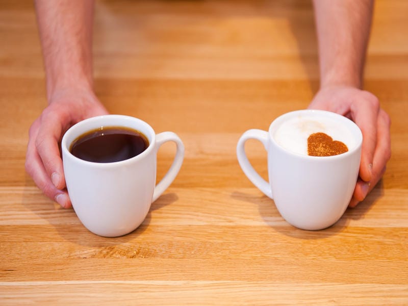 How Do Americano Vs Macchiato Differ in Caffeine Taste and Strength? 8 Two white coffee mugs held by hands, one with black Americano coffee and the other with Macchiato topped with milk foam and heart-shaped cinnamon