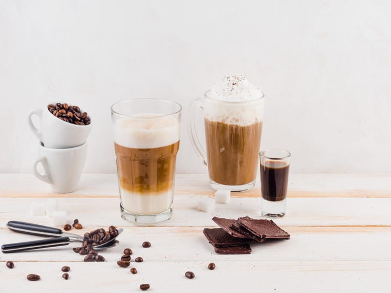 Various coffee drinks with chocolate milk, coffee beans, and chocolate pieces on a white wooden table