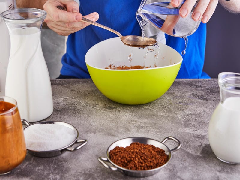 How Do You Make Mocha Drizzle in 7 Simple Steps? 7 Person pouring water into a bowl containing dry ingredients with milk and coffee powder on the table
