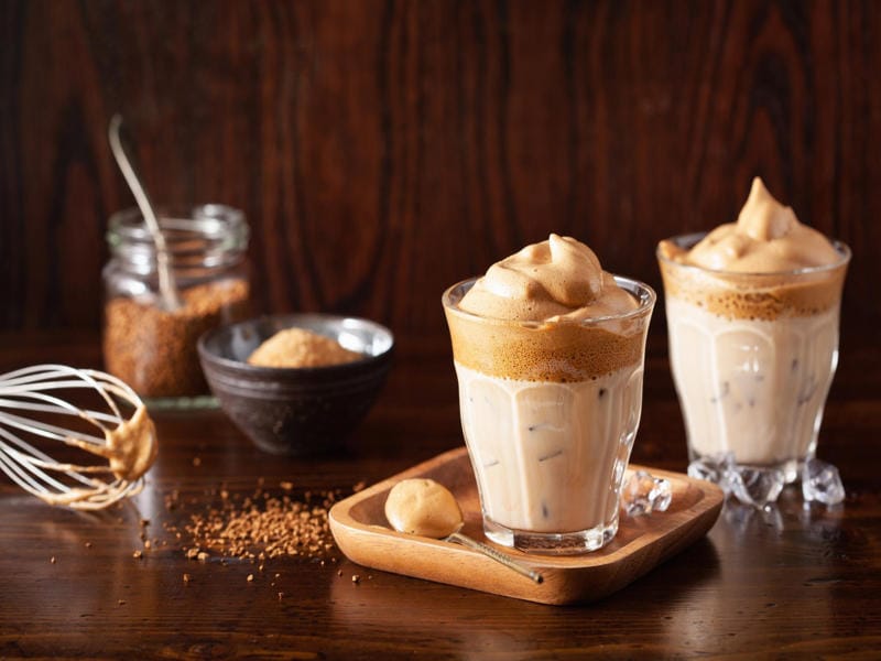 Two glasses of whipped coffee on ice with ingredients and whisk on wooden table, homemade Starbucks copycat recipe