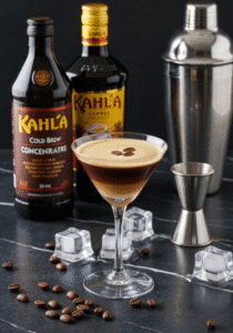 Elegant cold drip coffee cocktail in a martini glass garnished with espresso beans, surrounded by Kahlúa cold brew concentrate, coffee liqueur, ice cubes, a cocktail shaker, and a jigger on a dark marble surface.