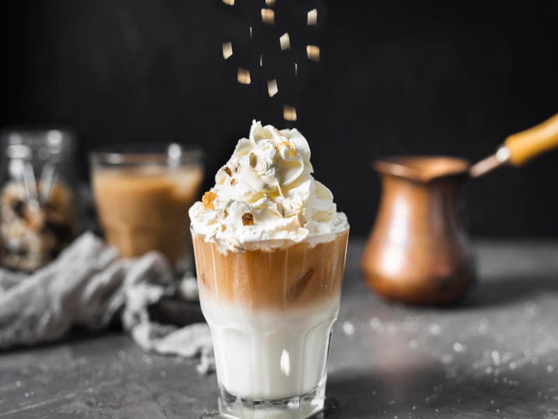 Close-up of white caramel mocha topped with whipped cream and caramel bits in a clear glass with coffee in the background