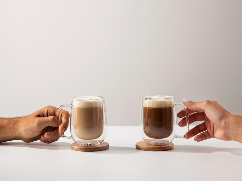 Two hands holding glass mugs of white mocha and mocha coffee on wooden coasters on white table